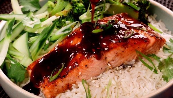 A bowl filled with rice topped with salmon, mixed green vegetables and a drizzle of teriyaki sauce