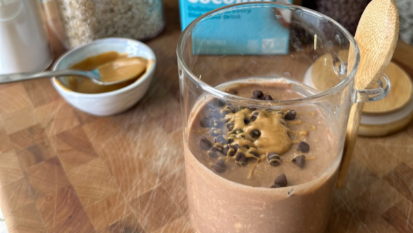 A small glass with a spoon attached and filled with peanut butter overnight oats topped with chocolate chips and peanut butter and sitting on top of wooden cutting board with a blue carton of silk milk behind it