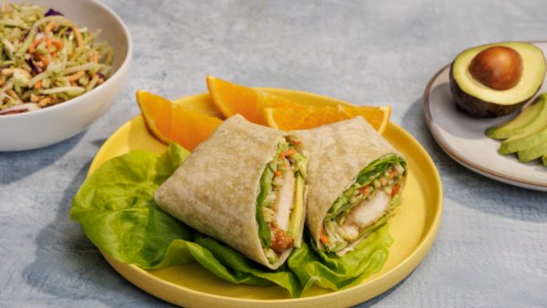 A halved avocado on a plate next to a spicy crunchy chicken wrap on a plate and a bowl of fried rice behind it